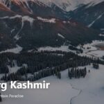 Panoramic view of Gulmarg in Kashmir, India, featuring snow-covered mountains, pine forests, and green valleys under a clear blue sky.